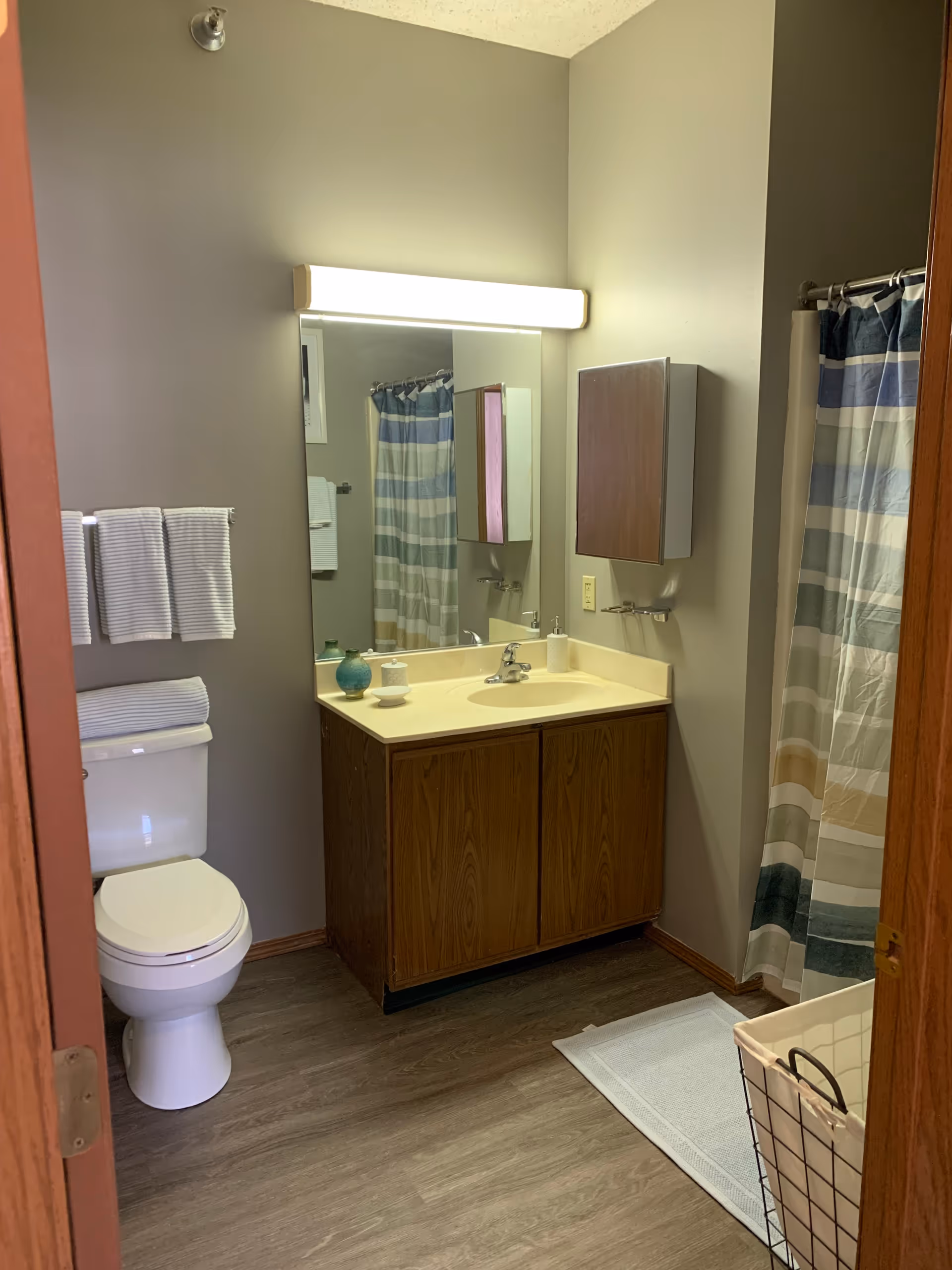 A bathroom with a white toilet, a wooden vanity with a sink, a large mirror above the sink, and a light fixture above the mirror. There are striped towels hanging on the wall and a striped shower curtain covering the bathtub. A white bath mat is on the floor next to a laundry basket.