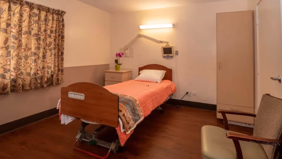 A simple bedroom with a single hospital-style bed covered with a coral blanket and a pillow. There is a small bedside table with a potted orchid, a wall-mounted medical monitor, a wooden wardrobe, and a cushioned chair. The room has wooden flooring and floral curtains covering the window.