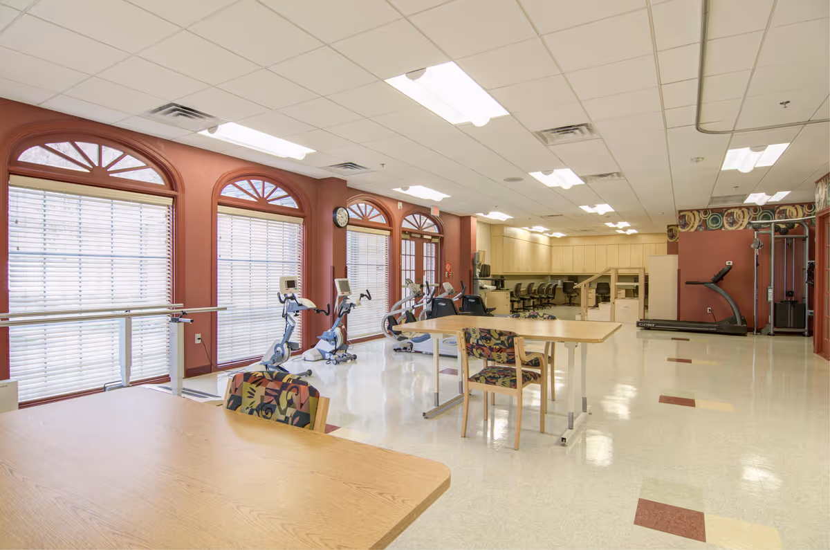 A spacious rehabilitation room with exercise equipment including stationary bikes and a treadmill, tables and chairs with patterned upholstery, large windows with blinds, and a counter with multiple chairs in the background.