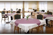 A bright dining room with multiple round tables covered with white tablecloths and purple table runners. Each table has a small vase with flowers. The room has large windows allowing natural light to fill the space, and wooden chairs with dark cushions are arranged around the tables.