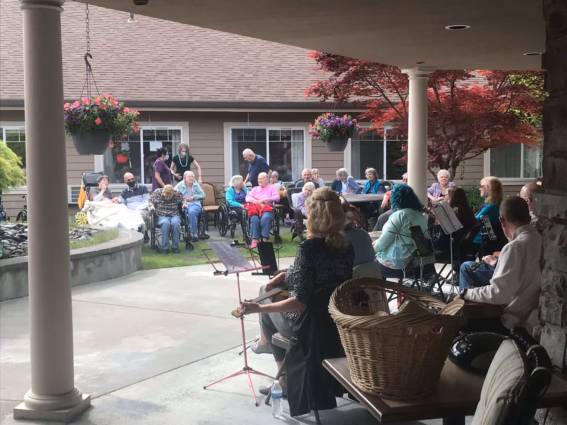 A group of elderly people sitting outside in wheelchairs and chairs, watching a small ensemble of musicians playing instruments under a covered patio. The setting is a garden area with hanging flower pots and a building with windows in the background.