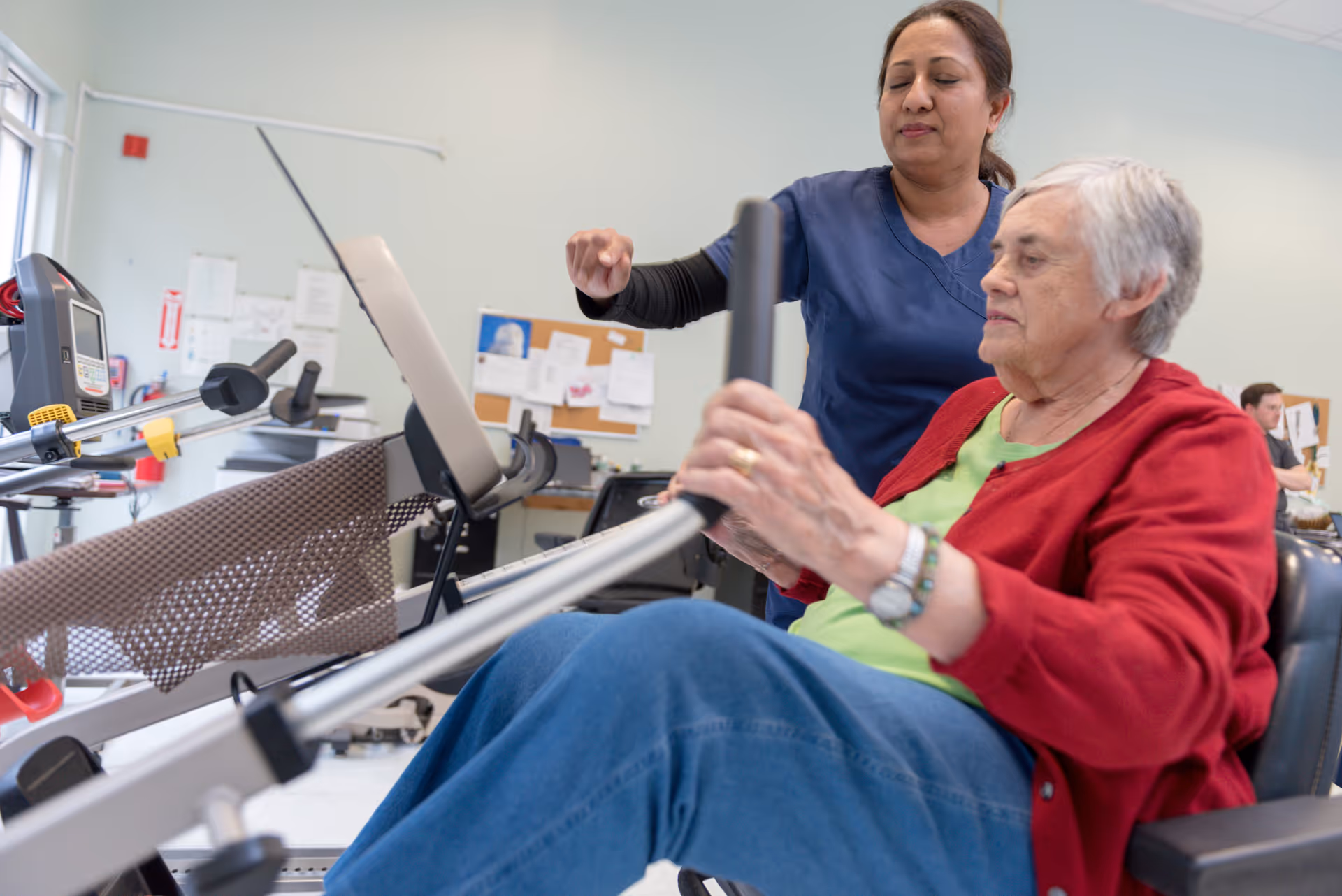 An elderly woman in a red cardigan and green shirt is using a rowing exercise machine in a rehabilitation center, assisted by a healthcare worker in blue scrubs. The setting appears to be a therapy or exercise room with equipment and a bulletin board in the background.