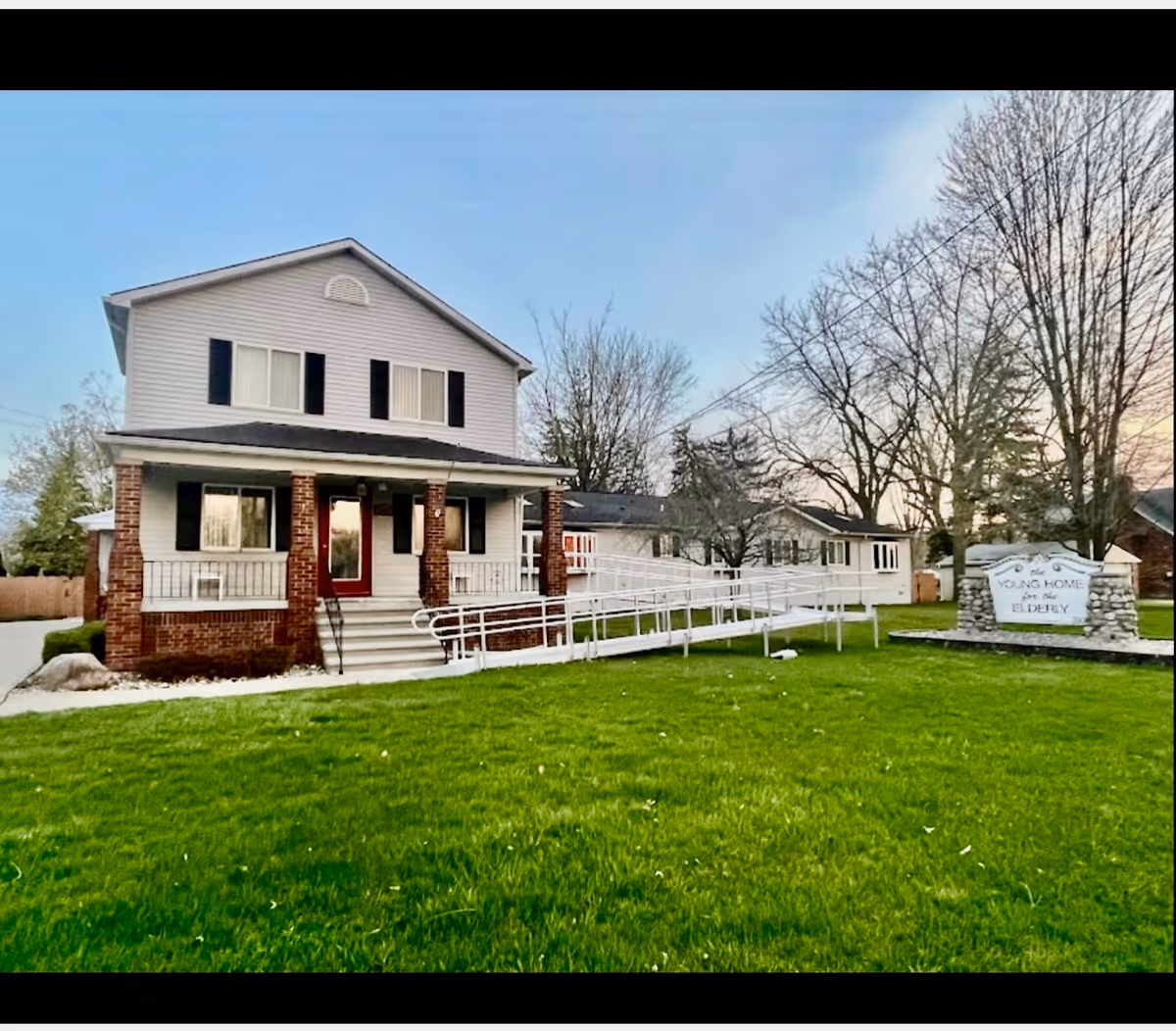 Two-story residential building with a front porch supported by brick columns, a wheelchair ramp leading to the entrance, and a well-maintained green lawn. To the right, there is a stone sign that reads 'The Young Home for the Elderly' with leafless trees in the background under a clear sky.