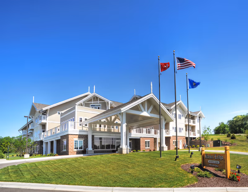 Three-story assisted living building with a covered porte-cochère, flagpoles, landscaped lawn and a wooden sign reading Glarner Lodge.
