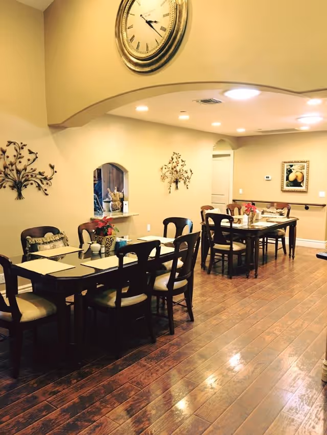 A dining room with two rectangular dark wood tables, each surrounded by six chairs with light-colored cushions. The tables are set with placemats, napkins, and small decorative items including a poinsettia plant. The room has wooden flooring, beige walls, decorative metal wall art, a large round clock above an archway, and a framed painting on the far wall. Recessed ceiling lights illuminate the space.