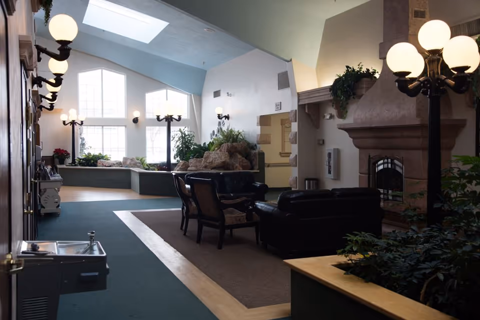 A spacious living room area with high ceilings and large windows allowing natural light to fill the space. The room features a stone fireplace, black leather sofas, wooden chairs, several plants, and multiple vintage-style lamp posts. There is a water fountain on the left side and a skylight above, creating a bright and inviting atmosphere.