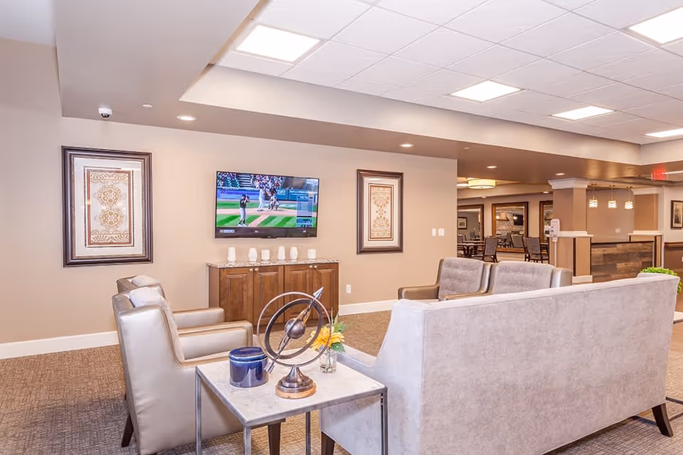 A bright and spacious living room area in an assisted living facility featuring beige walls and carpet. The room has a mounted flat-screen TV showing a baseball game, two framed decorative artworks on the wall, and comfortable seating including two armchairs and a sofa. A small side table holds decorative items including a metallic sculpture and a small plant. In the background, there is a dining area with tables and chairs and pendant lighting.