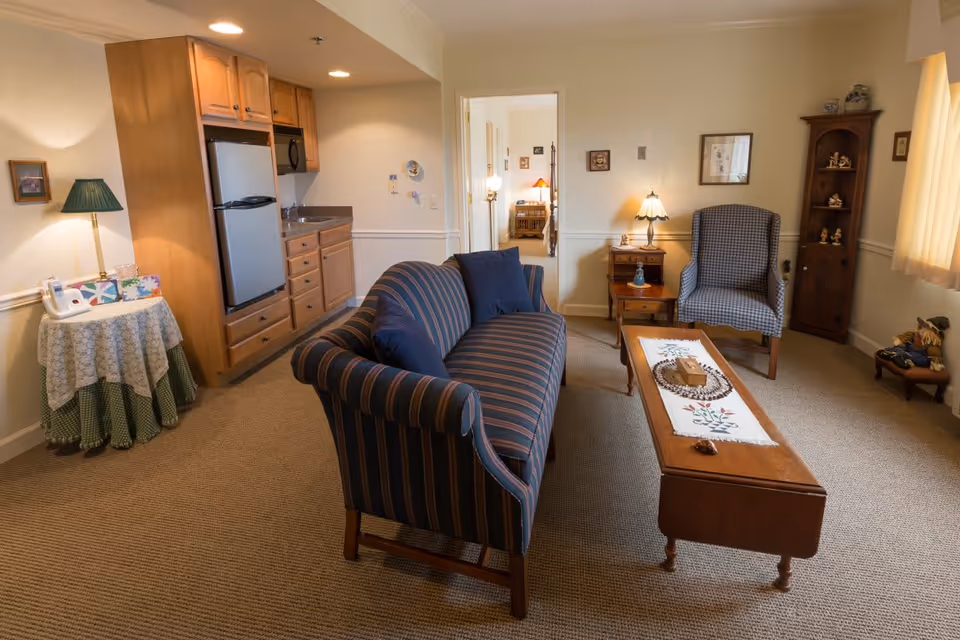 A cozy living area in a retirement community featuring a striped sofa with two blue pillows, a wooden coffee table with a decorative runner, a checkered armchair, and a small wooden corner shelf with figurines. To the left, there is a kitchenette with wooden cabinets, a small refrigerator, and a sink. A round table with a lace tablecloth and a green lamp is also visible. In the background, an open doorway leads to a bedroom with a bed and nightstand.