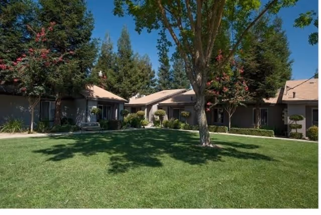 A sunny outdoor courtyard area with green grass, several trees, and a single-story building with beige walls and a tan roof surrounding the lawn.