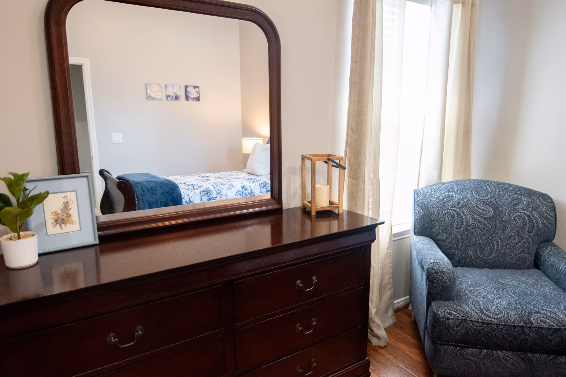 A cozy bedroom corner featuring a dark wooden dresser with a large mirror reflecting a bed with blue and white bedding. On the dresser are a small potted plant, a framed picture, and a decorative candle holder. Next to the dresser is a blue patterned armchair by a window with light-colored curtains.