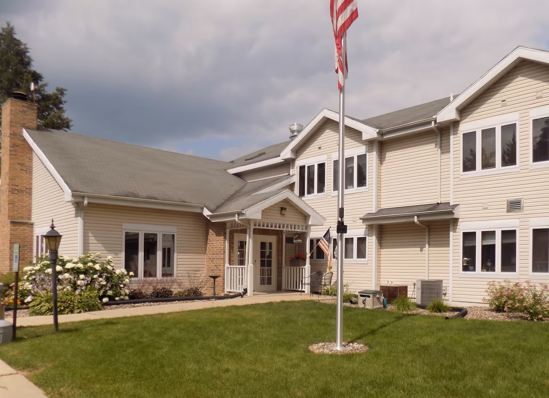 Two-story beige-siding senior living building with a front entrance, flagpole, and a grassy lawn.