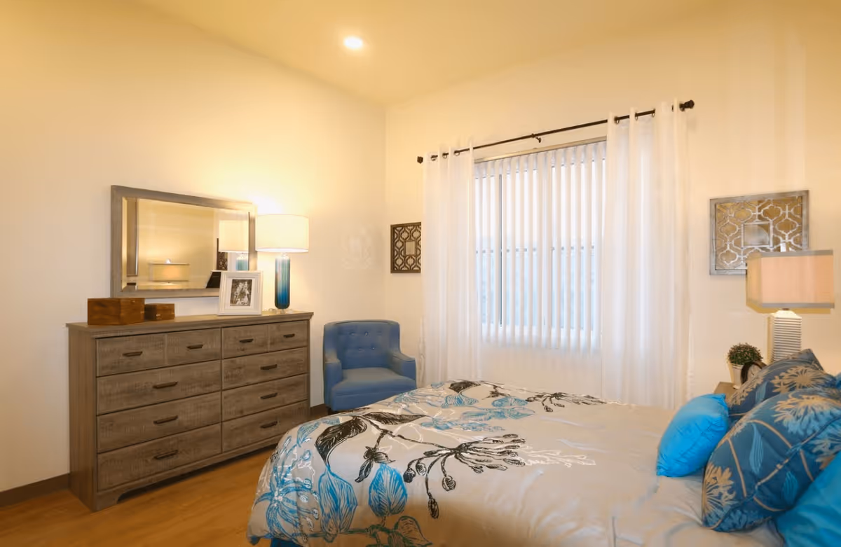 A cozy bedroom with a bed covered in a floral patterned comforter in shades of blue and beige. There are several blue and beige pillows on the bed. To the right of the bed is a nightstand with a lamp and a small plant. On the left side of the room is a wooden dresser with multiple drawers, a large mirror above it, a table lamp, and a framed photo. A blue upholstered chair is placed in the corner near a window with white vertical blinds and sheer white curtains. The room has light-colored walls and wooden flooring.