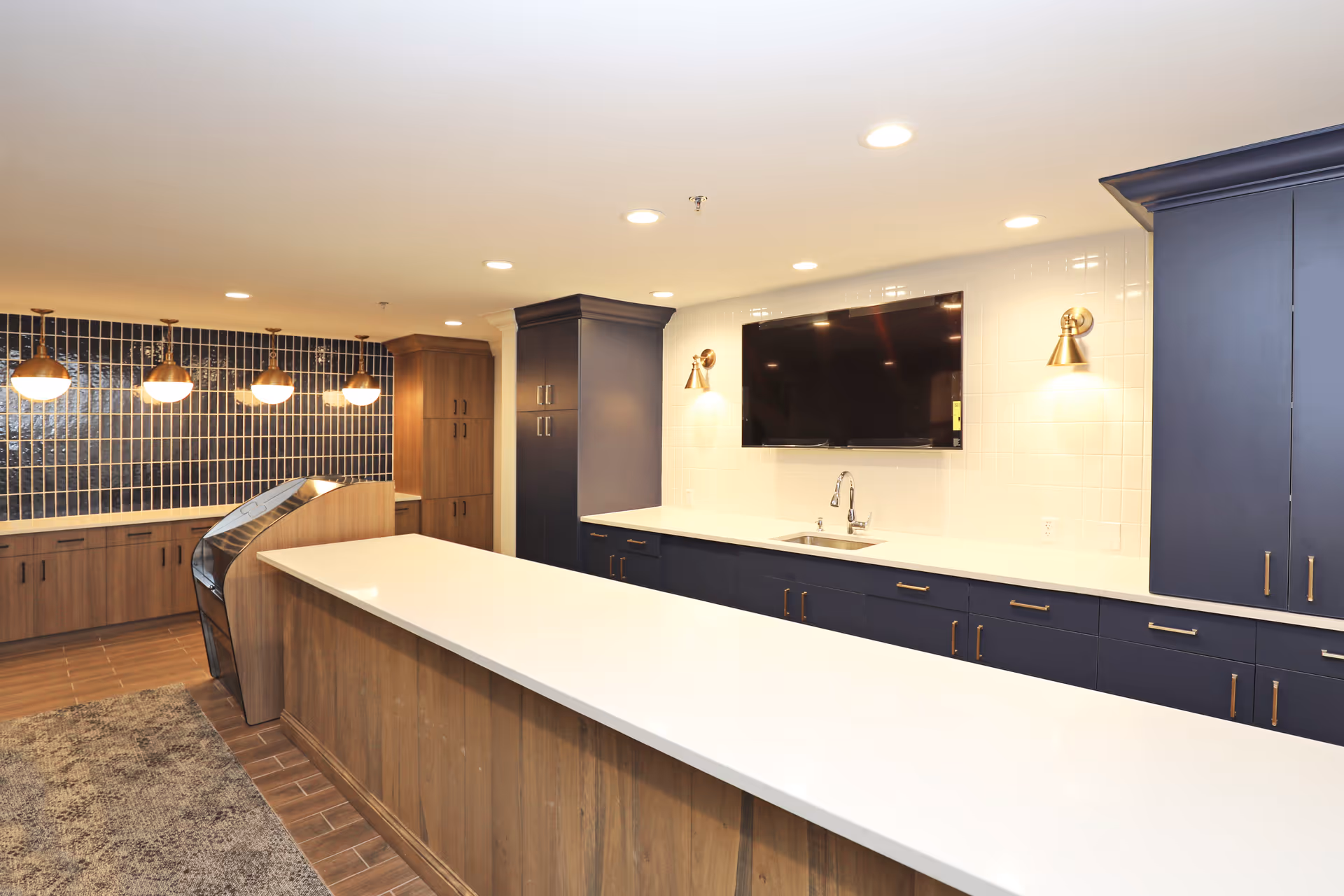A modern kitchen area with a long white countertop island in the foreground, dark blue lower cabinets with brass handles, a sink under a wall-mounted flat-screen TV, and a backsplash of white tiles. On the left side, there are wooden cabinets and a wall with vertical dark blue tiles, illuminated by four pendant lights hanging from the ceiling.