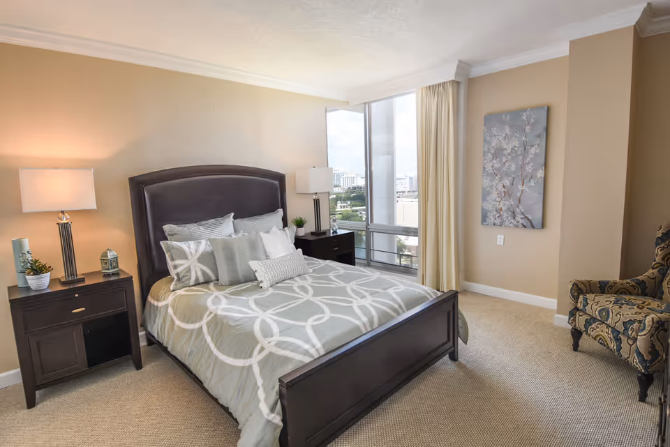 A well-lit bedroom with a large bed featuring a patterned gray and white comforter and multiple pillows. There are two dark wooden nightstands on either side of the bed, each with a lamp and decorative items. A large window with beige curtains offers a view of buildings outside. A patterned armchair is positioned in the corner, and a floral painting hangs on the wall.