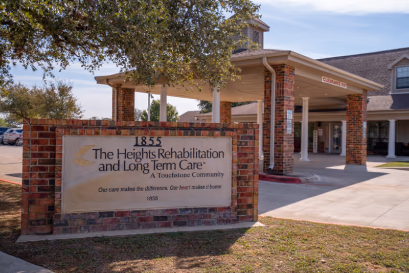 Entrance of The Heights Rehabilitation and Long Term Care facility showing a brick sign with the facility name and slogan, and a covered driveway with brick pillars and a clearance sign.