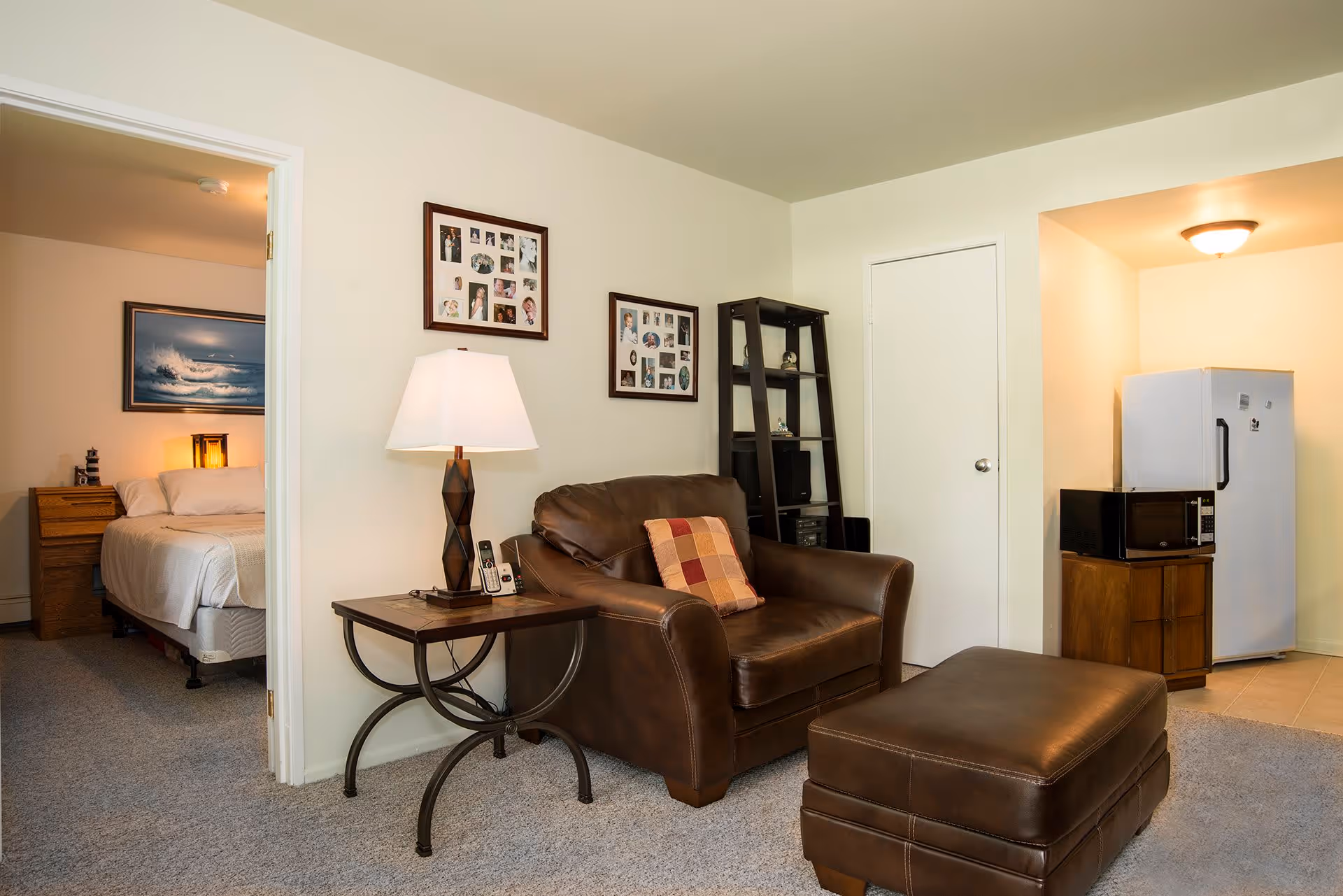 A cozy living room area with a brown leather armchair and matching ottoman. Next to the armchair is a side table with a lamp and a cordless phone. On the wall above the armchair are two picture frames with multiple photos. To the right, there is a small kitchen area with a white refrigerator and a microwave on a wooden stand. To the left, an open door reveals a bedroom with a bed, nightstand, and a painting of ocean waves on the wall.