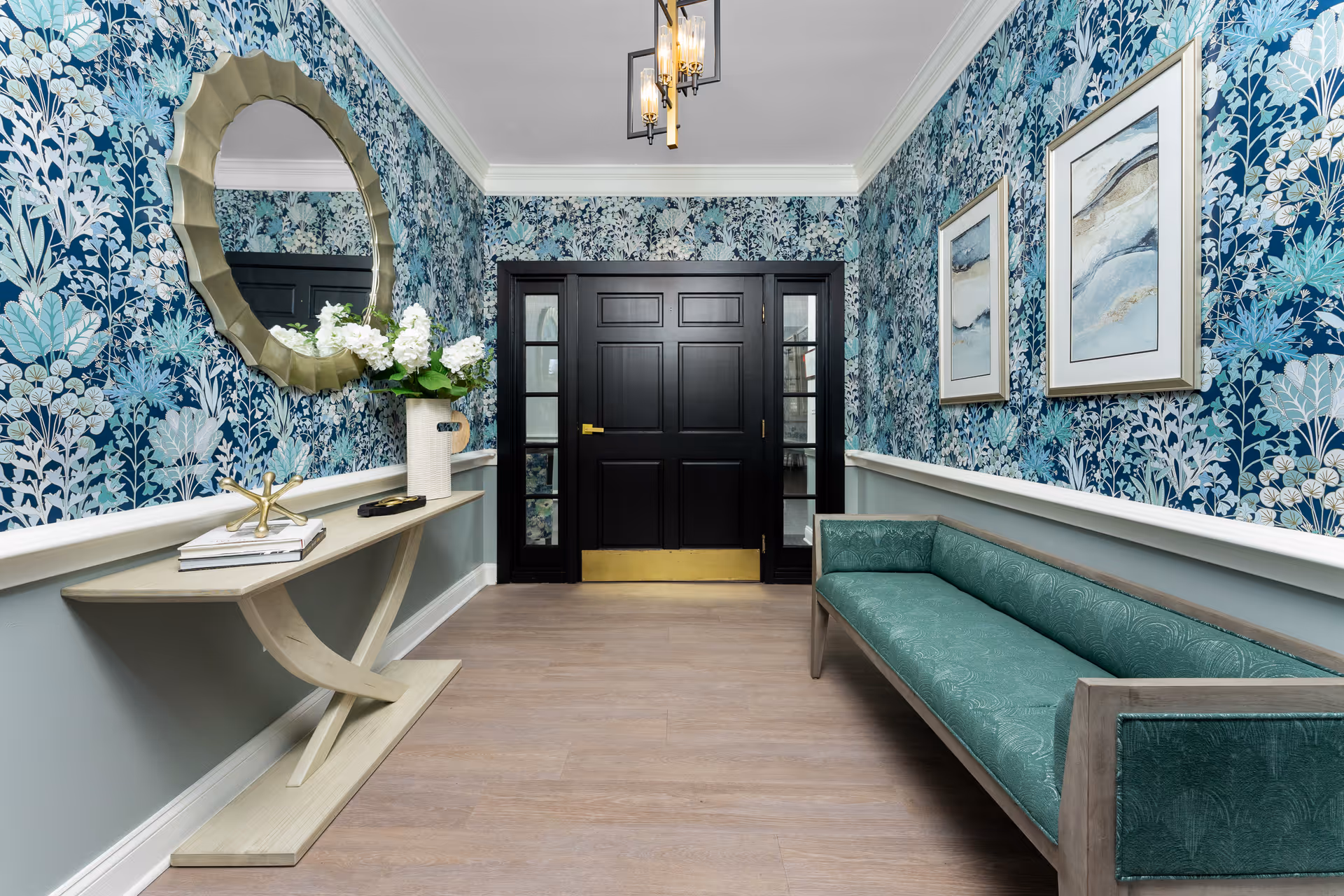 A hallway with floral blue wallpaper, a wooden floor, and a black double door at the end. On the left side, there is a narrow console table with a decorative gold sculpture, books, and a white vase with white flowers beneath a round gold-framed mirror. On the right side, there is a green upholstered bench and two framed abstract paintings on the wall. A modern chandelier hangs from the ceiling.