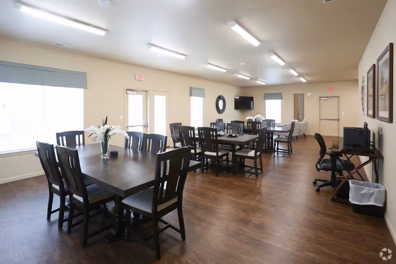 A spacious, well-lit dining room with multiple dark wooden tables and chairs arranged neatly. There are large windows with light blue valances allowing natural light to fill the room. A wall-mounted TV and decorative items like a round mirror and framed pictures are visible. A desk with a computer and office chair is positioned against one wall, and the floor is covered with wood-style flooring.