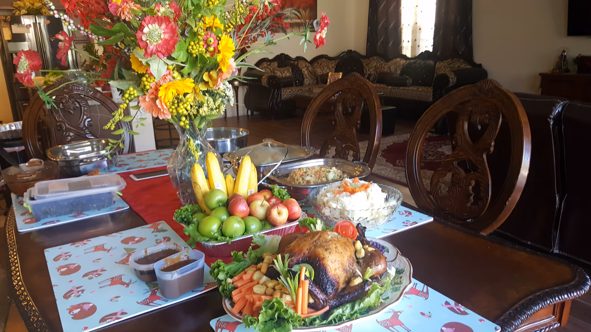 A dining table set with a variety of food including a roasted chicken with vegetables, a bowl of fruit with bananas and apples, a bowl of salad, and covered dishes. A large vase with colorful flowers is also on the table. In the background, there is a living room area with ornate wooden chairs, a sofa, and a window with curtains.