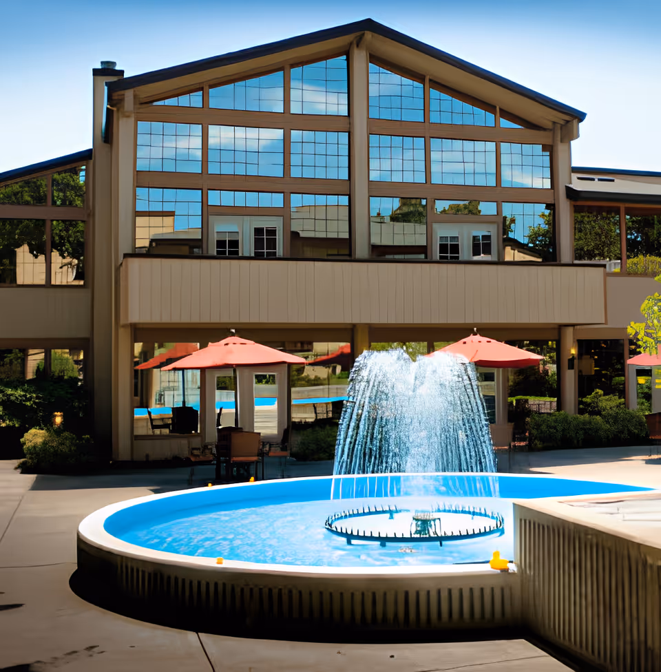 Outdoor courtyard area of Oakmont Gardens featuring a round water fountain with water spraying upwards, surrounded by a paved area. In the background, there is a building with large reflective windows and red patio umbrellas providing shade over seating areas.