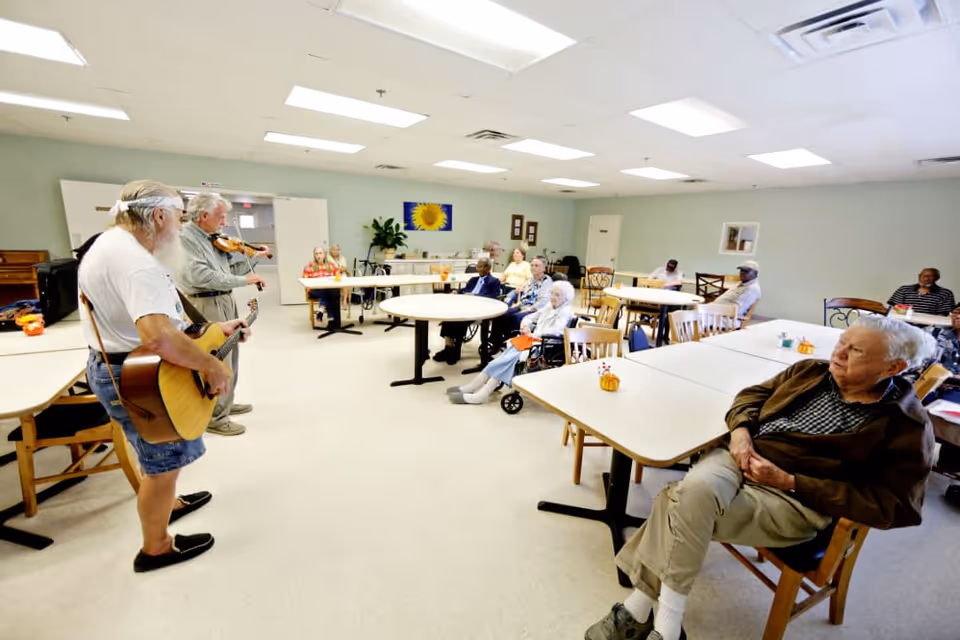 Residents sit at tables in a communal dining/activity room while two musicians play guitar and violin.