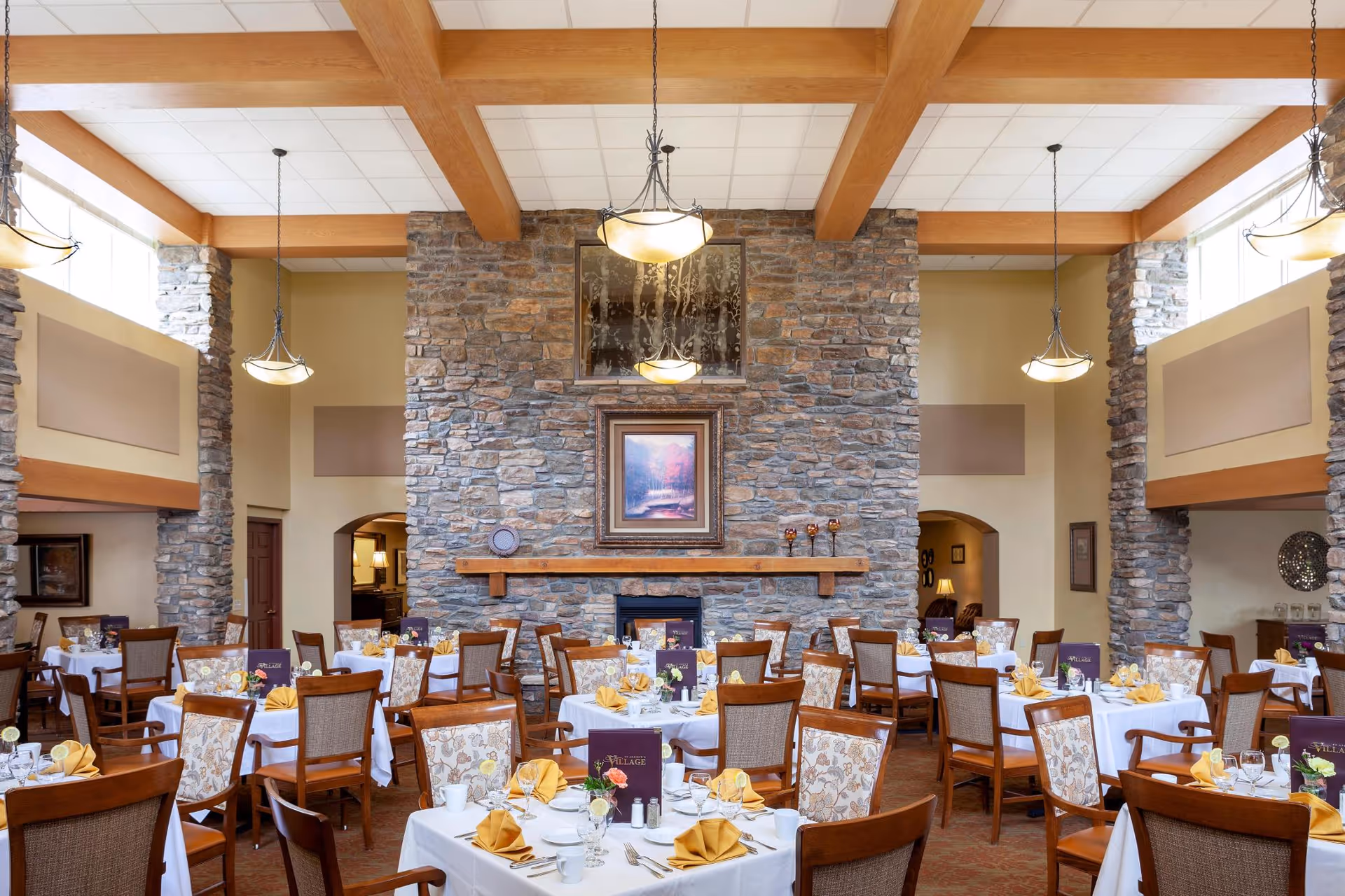 A spacious dining room with multiple tables set for a meal, featuring white tablecloths, yellow folded napkins, glassware, and floral centerpieces. The room has high ceilings with wooden beams, stone pillars, and a large stone fireplace with a framed painting above it. Pendant lights hang from the ceiling, and there are arched doorways and large windows allowing natural light.
