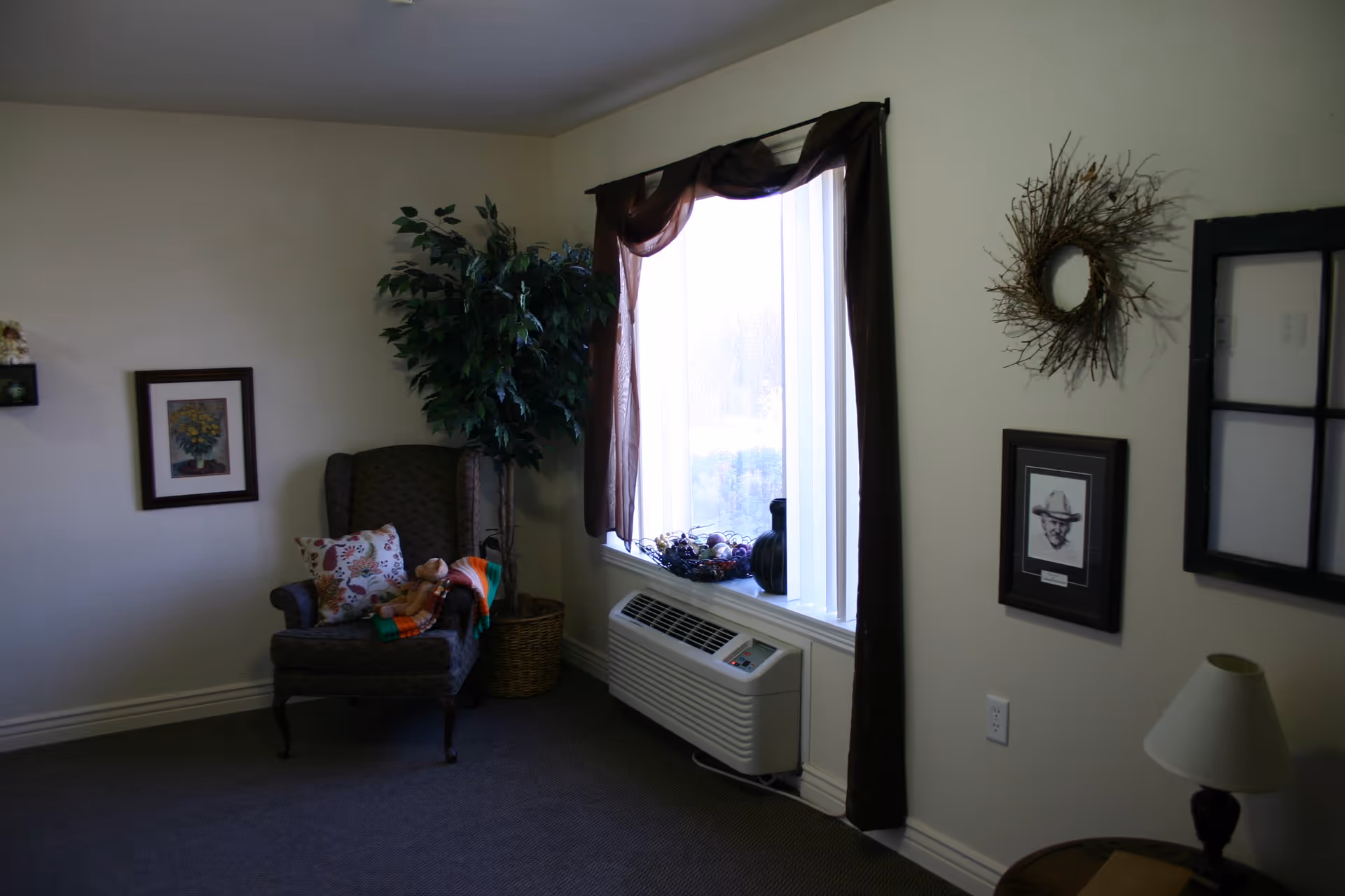 A cozy corner of a living room with a patterned armchair holding a floral pillow and a teddy bear with a colorful blanket. Next to the chair is a tall potted artificial plant. A window with sheer white curtains and dark brown drapes lets in natural light, with decorative items on the windowsill. The walls are decorated with framed pictures and a wreath made of twigs. A small table with a lamp is partially visible.