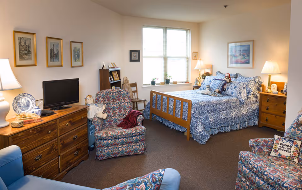 A cozy bedroom in a senior living facility with a wooden bed dressed in blue and white patterned bedding. The room features two patterned armchairs, a wooden dresser with a small TV, framed artwork on the walls, and a window letting in natural light. There are lamps on the dresser and bedside tables, along with decorative items and dolls placed around the room.