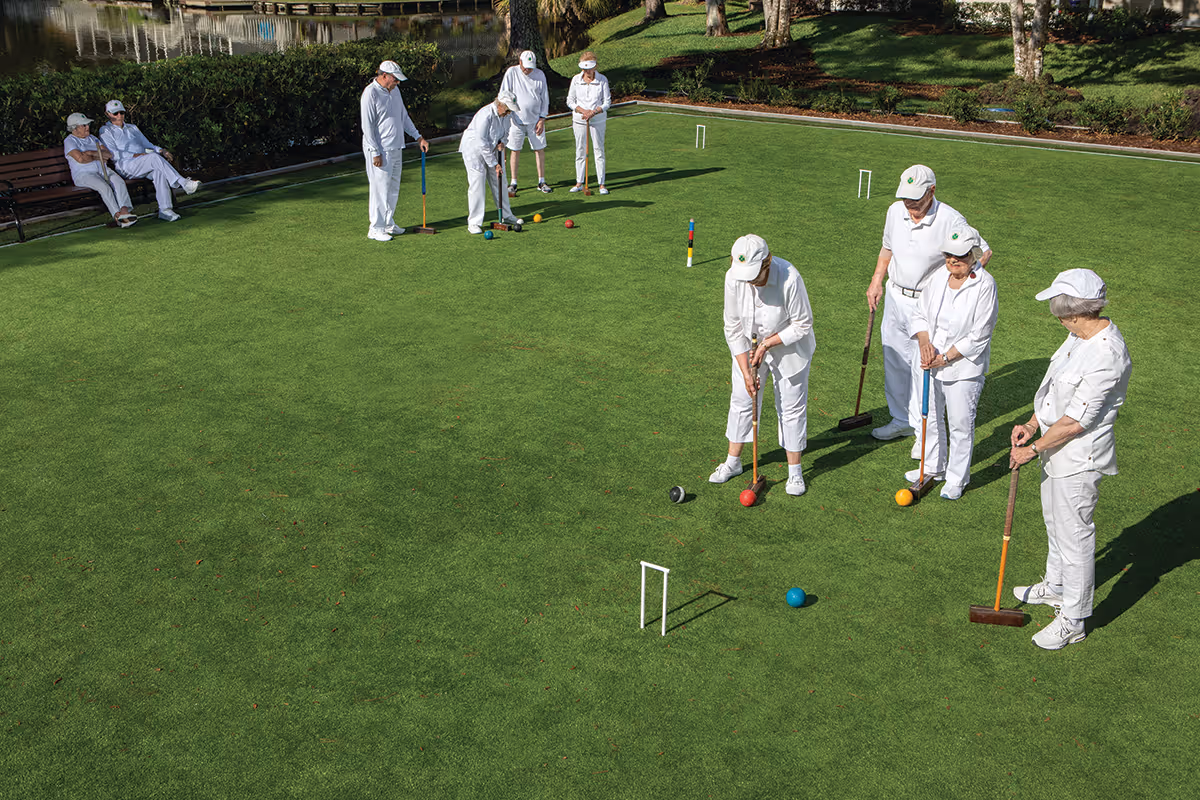 A group of elderly people dressed in white are playing croquet on a well-maintained green lawn. Some are actively playing while others are sitting on a bench nearby. The setting is outdoors with trees and a body of water in the background.