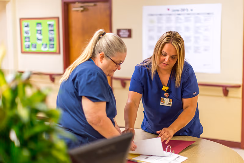 Two healthcare workers in blue scrubs reviewing documents together at a round table in a healthcare facility hallway.
