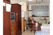 Nursing home bedroom with a wooden wardrobe and dresser holding an old TV, a wheelchair, small table, and a bed by a window.