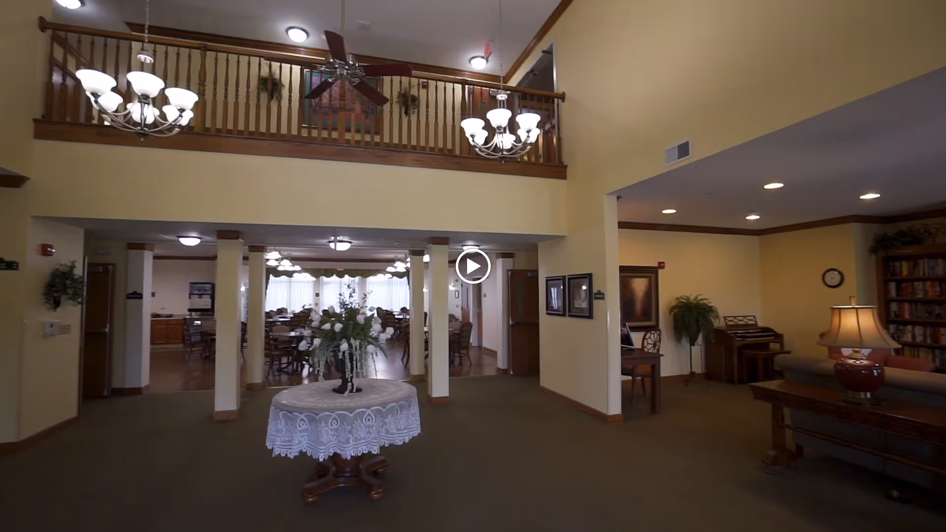 Interior view of a senior living facility showing a spacious common area with a round table covered with a lace tablecloth and a floral arrangement in the center. To the left, there is a dining area with multiple tables and chairs under hanging light fixtures. On the right, there is a cozy sitting area with a lamp, a piano, and a bookshelf. The room has high ceilings with a wooden balcony railing and ceiling fans.