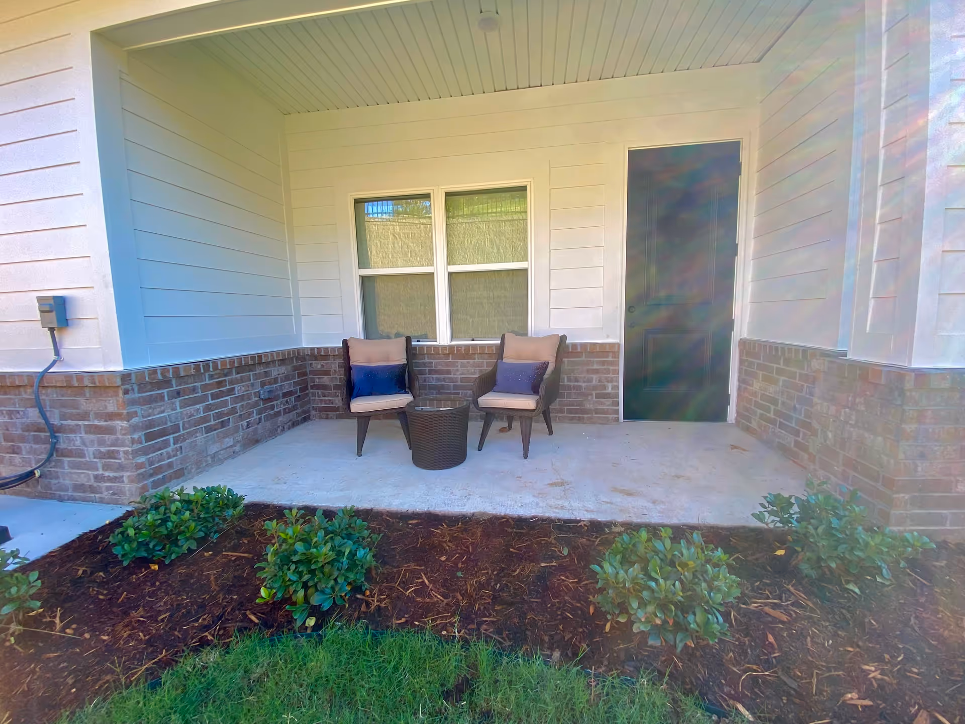 A small outdoor patio area with two cushioned chairs and a small round table between them. The patio is adjacent to a building with white siding and brick lower walls. There is a window and a closed door on the wall behind the chairs. In front of the patio, there are small green bushes and a patch of grass.