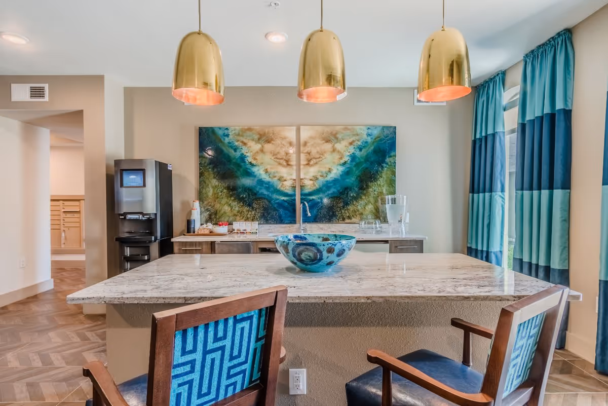 Interior view of a modern common area with a marble countertop island, two wooden chairs with blue patterned cushions, three gold pendant lights hanging from the ceiling, a decorative blue bowl on the countertop, a water dispenser on the left, and a large abstract painting with blue and green tones on the wall behind the counter. Blue and teal curtains cover the windows on the right side.