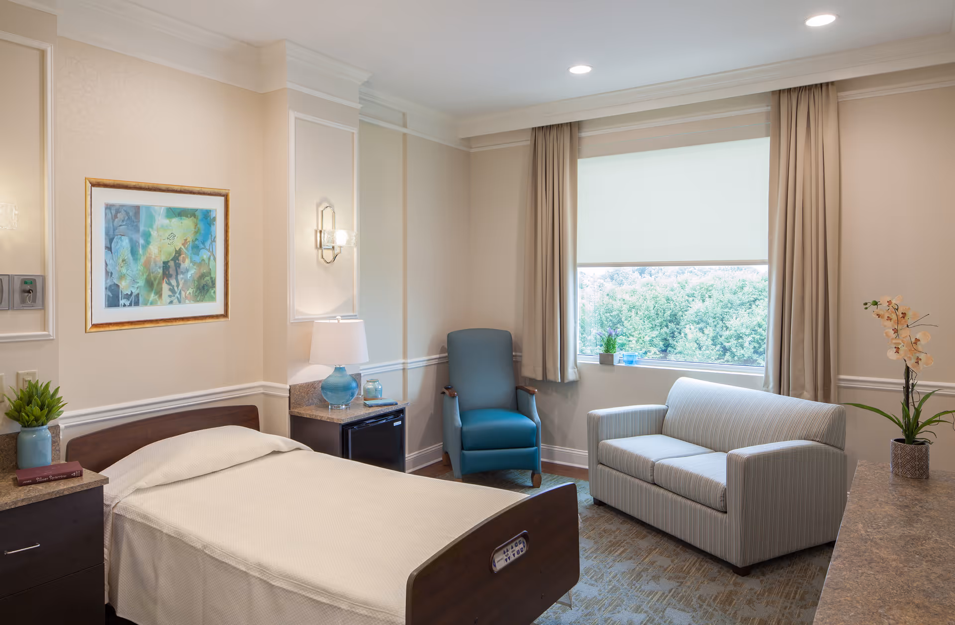 A well-lit bedroom in a senior living facility featuring a single bed with white bedding, a wooden headboard, a nightstand with a blue lamp and a small plant, a comfortable blue armchair, a striped loveseat, and a large window with beige curtains overlooking greenery outside. The walls are light-colored with decorative molding and a framed abstract painting.