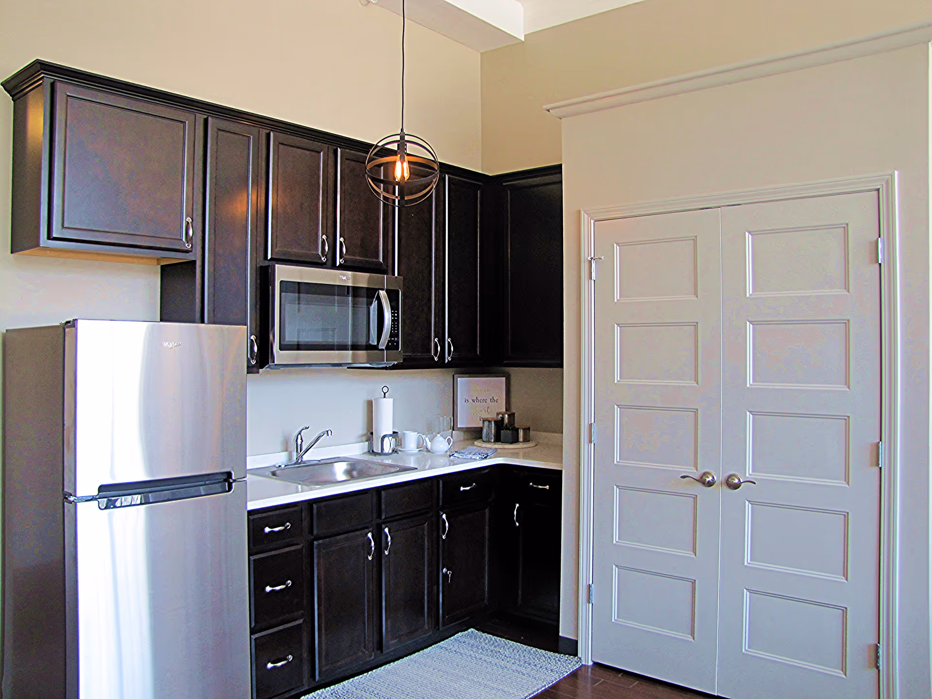 Small kitchen with dark wood cabinets, stainless steel refrigerator and microwave, a sink on a white countertop, and double pantry doors.