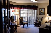 A cozy living room area with two wooden chairs, a small table with a lamp, a television on a stand, and vertical blinds covering a sliding glass door leading to an outdoor space. The room has carpeted flooring, a framed picture on the wall, and a window with a valance curtain.