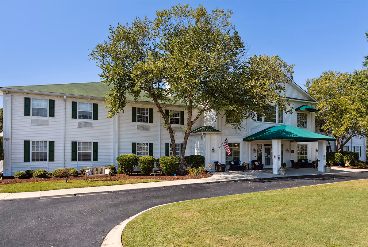 Exterior view of a two-story white building with green shutters and a green roof. There is a covered entrance with a green awning and outdoor seating. Trees and shrubs surround the building, and a paved driveway curves in front of the entrance under a clear blue sky.