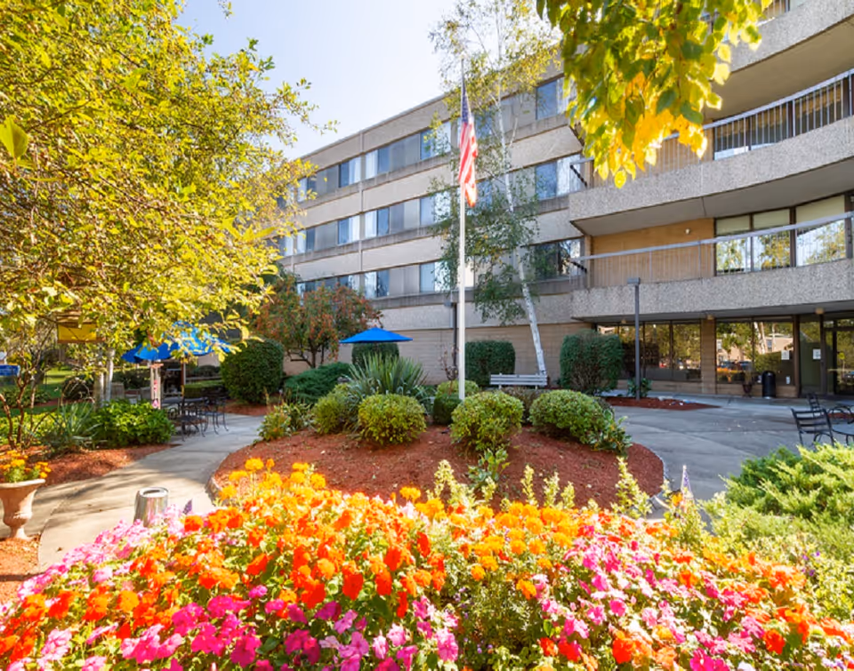 Courtyard garden with bright flowers, tables with blue umbrellas, an American flag, and a multi-story health and rehabilitation building in the background.