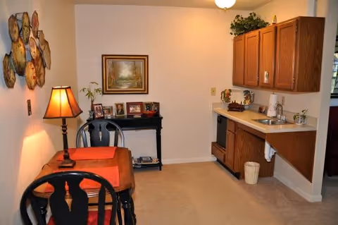 A small dining and kitchenette area with a wooden table and two chairs, a table lamp, a small black console table with framed photos, a painting on the wall, and wooden cabinets above a countertop with a sink and a small dishwasher. The room is carpeted and warmly lit.