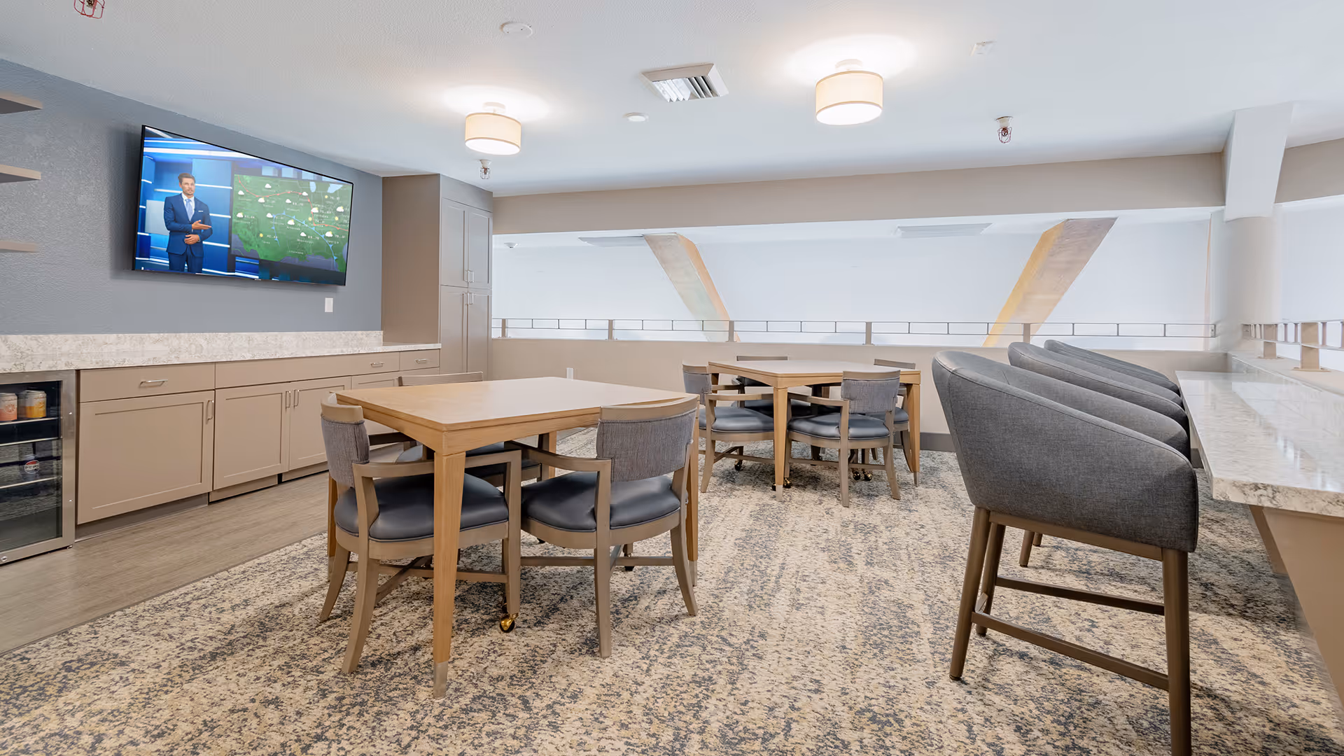 A modern common area with two wooden tables surrounded by chairs, a row of high-back chairs along a marble countertop, a flat-screen TV mounted on a gray wall displaying a weather forecast, and light-colored cabinetry with a small refrigerator underneath.