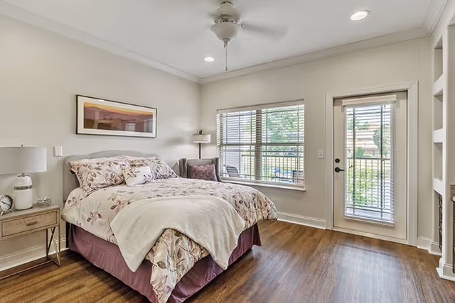 Sunlit bedroom with a floral-covered bed, nightstand, ceiling fan, large window and a glass door opening to a balcony.
