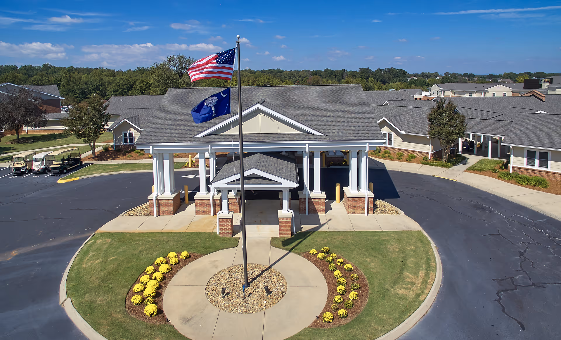 Front exterior view of a senior living facility named Rolling Green Village with a covered entrance, two flagpoles displaying the American flag and a state flag, surrounded by a circular driveway and landscaped with yellow flowers and green grass.