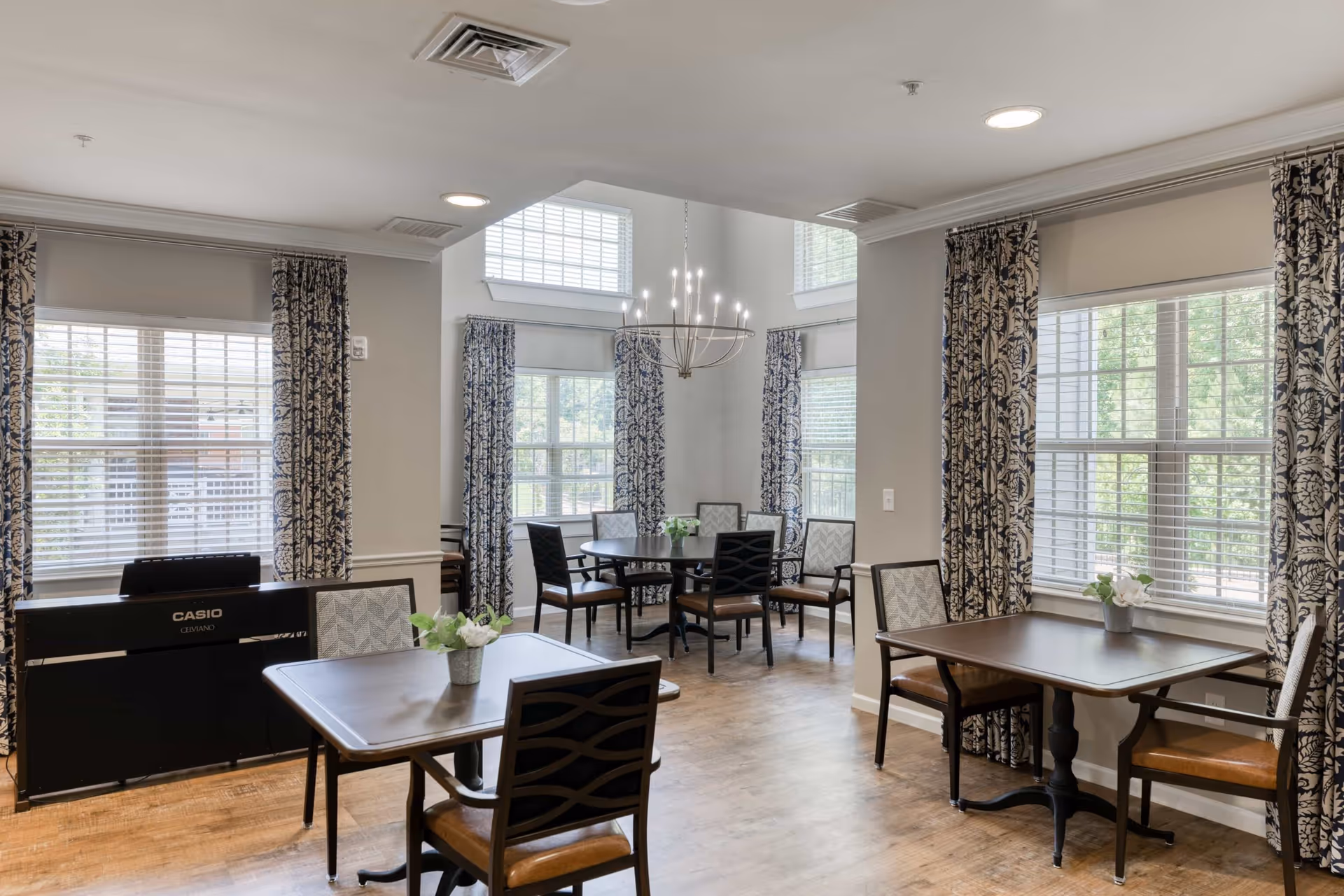 Bright communal dining room with multiple tables and chairs, large windows with patterned curtains, and a chandelier.