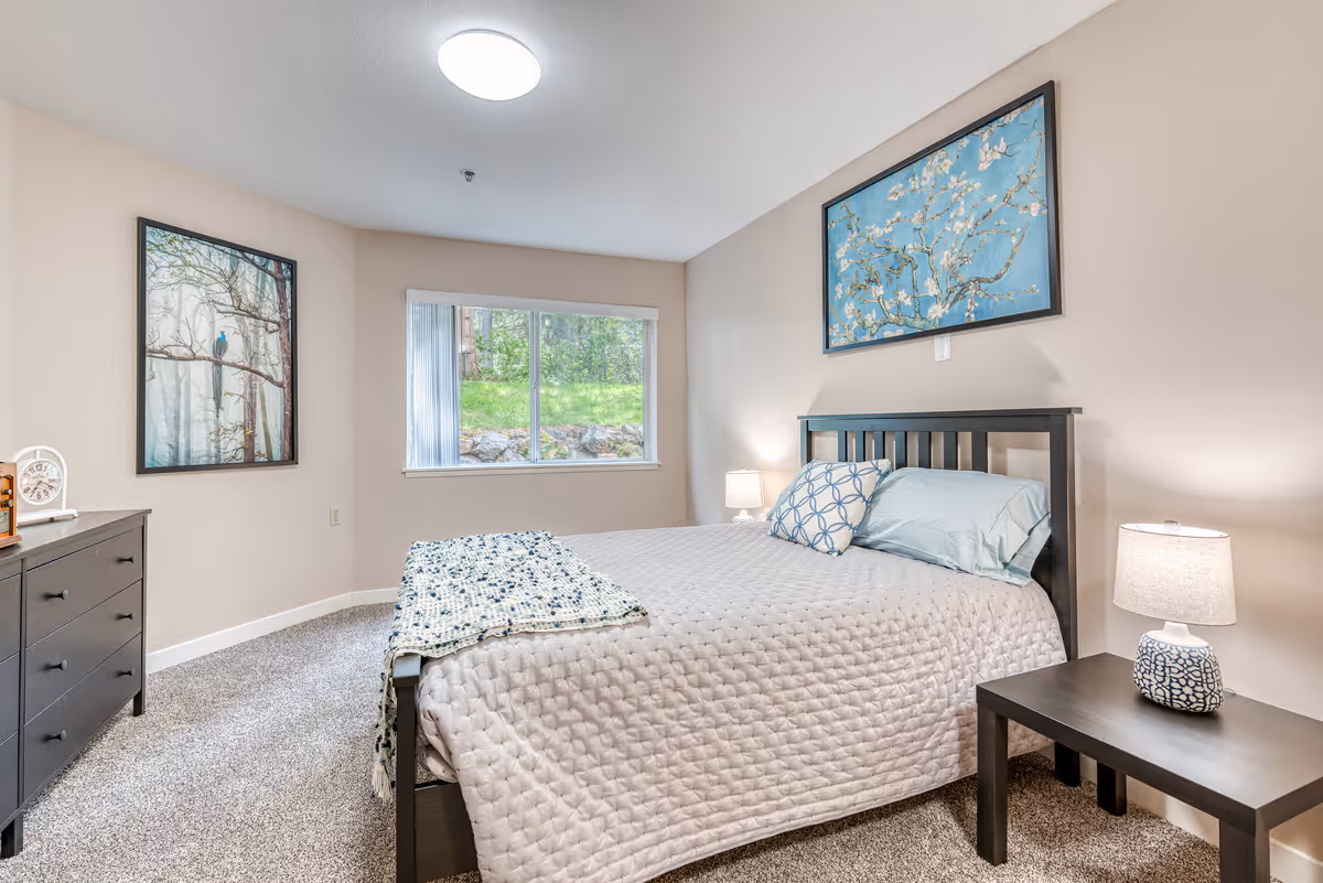 A neatly furnished bedroom with a bed, two nightstands with lamps, a dresser, a window, and framed artwork on the walls.