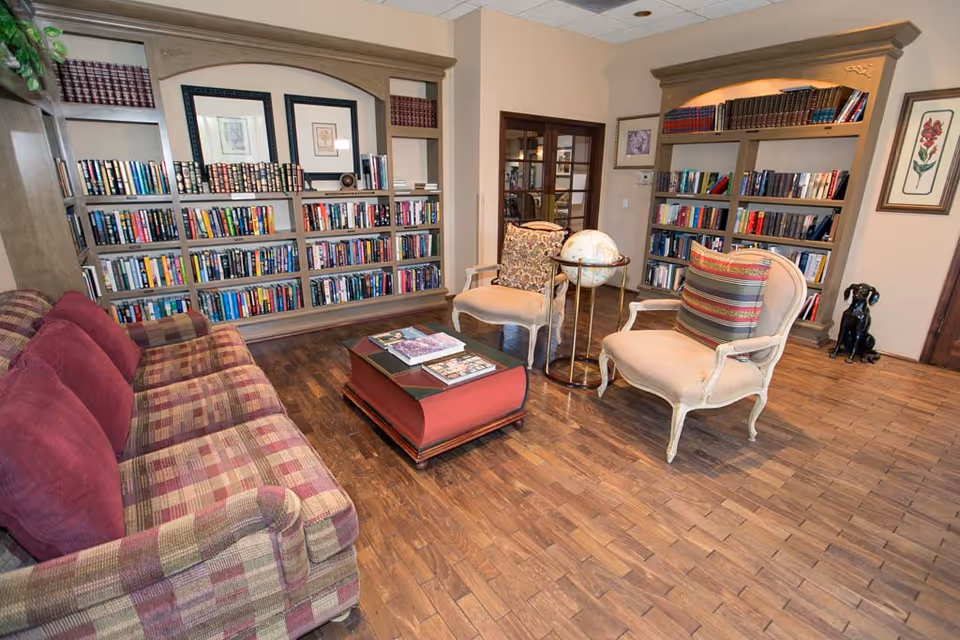 A cozy library or reading room with wooden floors, a plaid sofa with red cushions, two upholstered armchairs with patterned pillows, a coffee table shaped like a large book with magazines on top, and built-in bookshelves filled with books. There is also a globe on a stand and framed artwork on the walls.