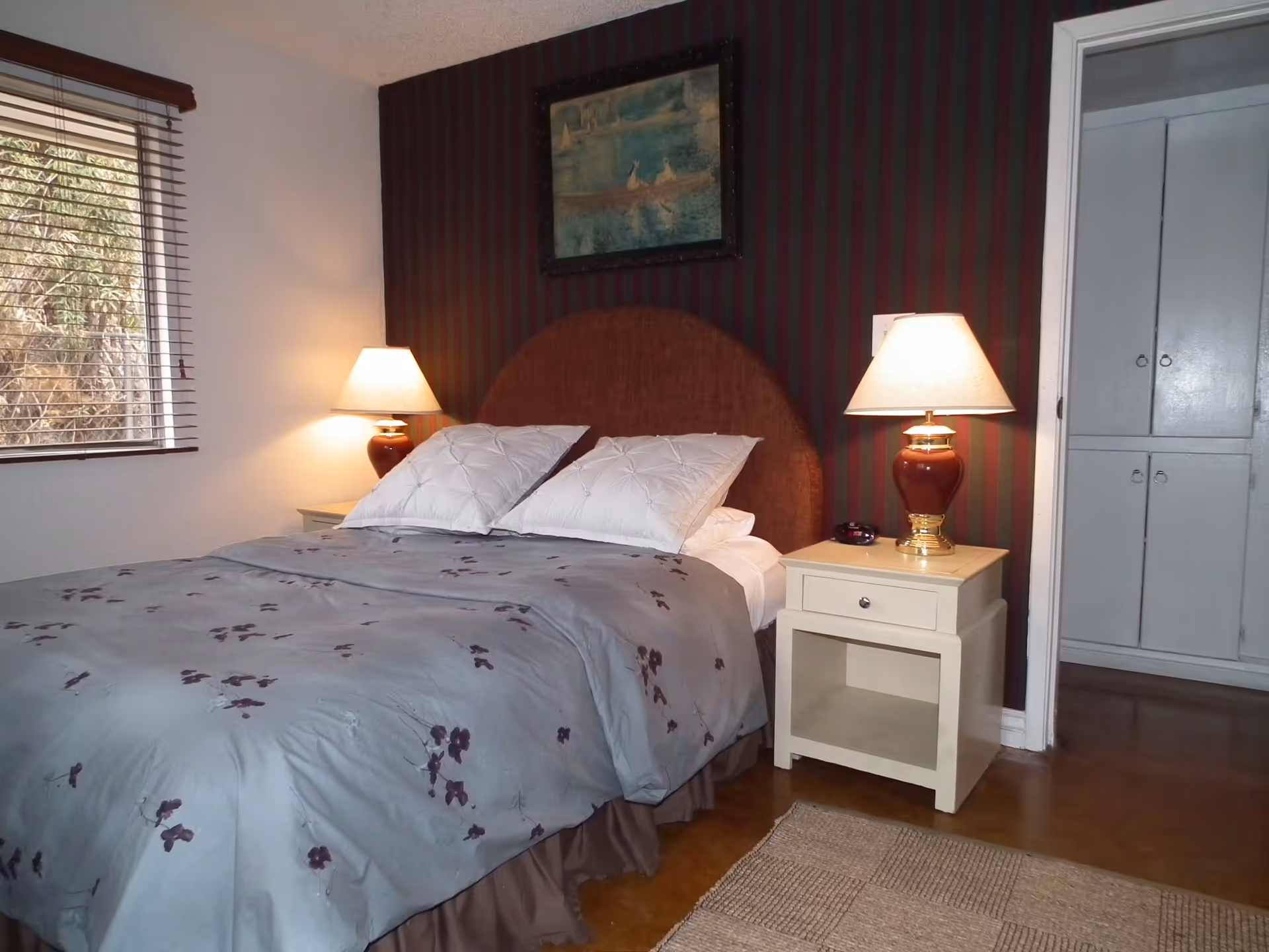 A bedroom with a bed covered in a gray floral comforter and two white pillows. There are two bedside tables, each with a lamp with a red base and beige shade. A window with wooden blinds is on the left wall, and a painting hangs above the bed on a dark striped accent wall. A doorway on the right leads to a closet with white double doors.