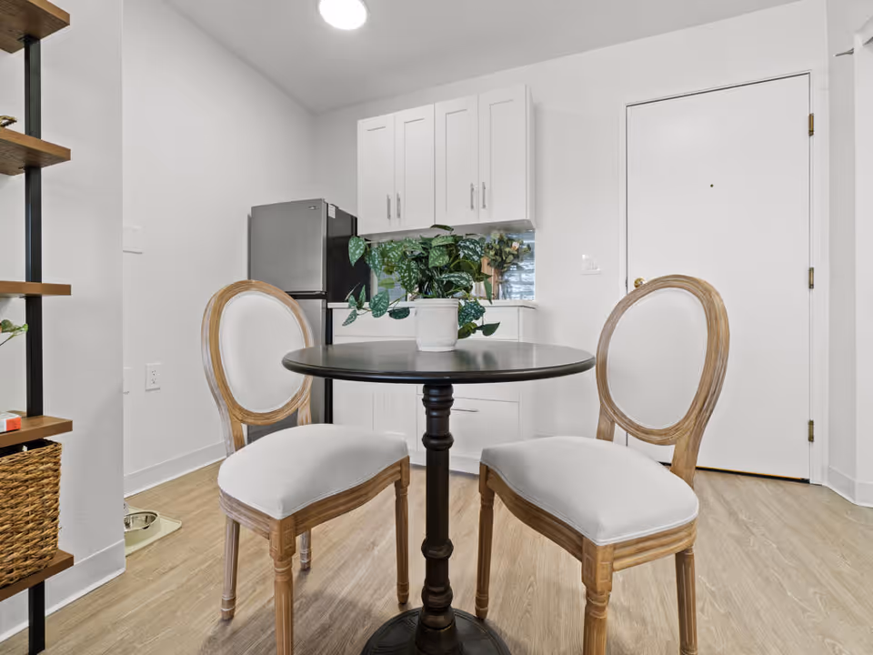 A small dining area with a round black table and two wooden chairs with white cushions. Behind the table is a compact kitchen area with white cabinets, a small refrigerator, and a potted plant on the table. The floor is light wood, and there is a white door to the right.