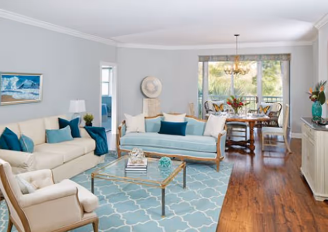 Bright open living and dining area with teal-accent sofas, a glass coffee table on a patterned rug, and a dining table by sliding glass doors.