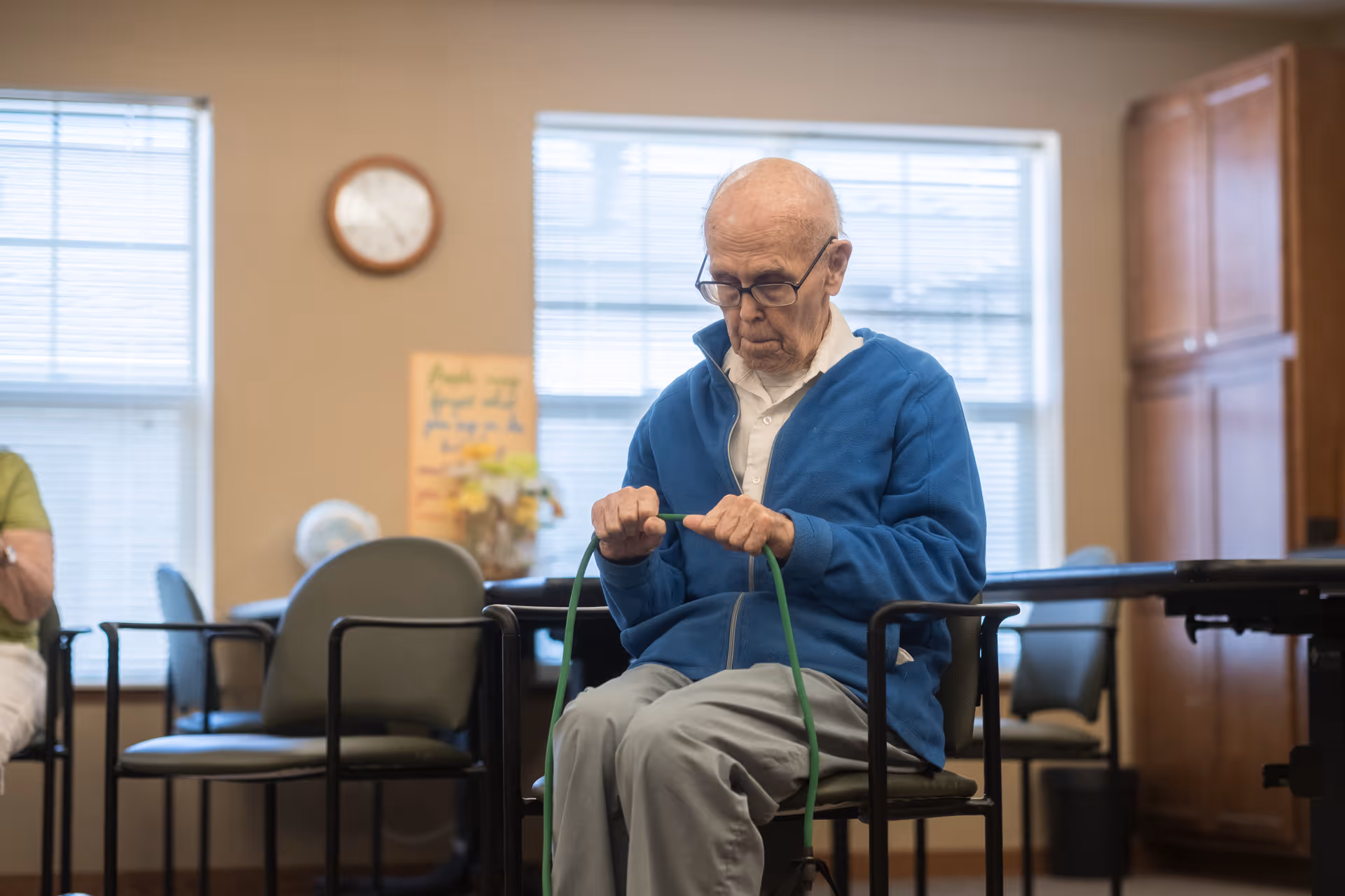 An elderly man wearing glasses and a blue jacket is seated on a chair in a room with large windows. He is holding a green exercise band and appears to be focused on it. The room has several chairs, a clock on the wall, and wooden cabinets in the background.